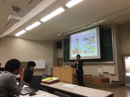 講義室の前方に立つ男性がスクリーンを使って説明を行い、手前では数名が席に着いて話を聞いている写真
