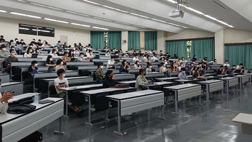 段々状に並んだ机がある教室で学生たちが座って講義を受けている写真