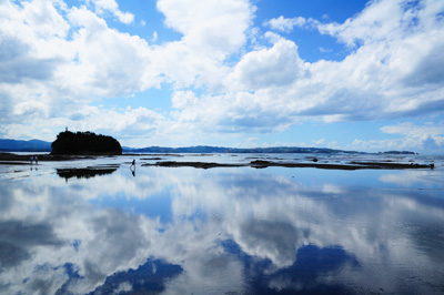 雲のかかった空が反射している海に3人の小さな人影が写っている写真
