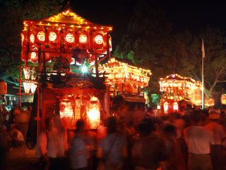 夜の暗い空で、飾りがつけられて明かりが灯された3台の山車と、山車を囲んでたくさんの人たちが写っている写真