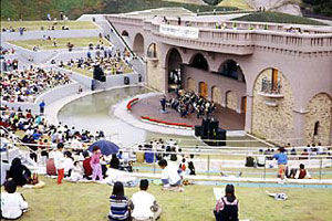 階段状の芝生席に大勢の観客が集まり、野外コンサートを楽しんでいるミューズパーク・たなべの写真