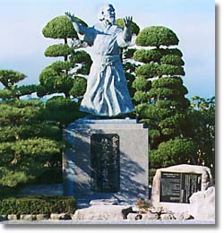 手入れされた木々の間に設置された胴着と袴を身に着けた男性が両手を広げて立っている銅像の写真