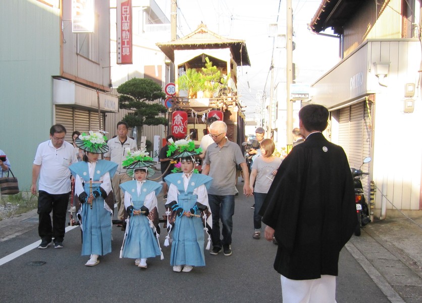 水色の和服に花の付いた笠を身に着けた3人の女の子が山車の前を歩いている写真