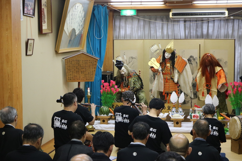 屏風を背にして立つお面や鎧を身に着けた人形の前に座って笛などを演奏する人たちの写真
