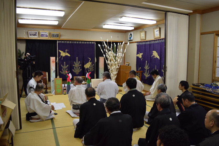 お供え物の最前列に座る白い和服を着た男性たちとその後ろに座る黒い和服を着た人たちの写真