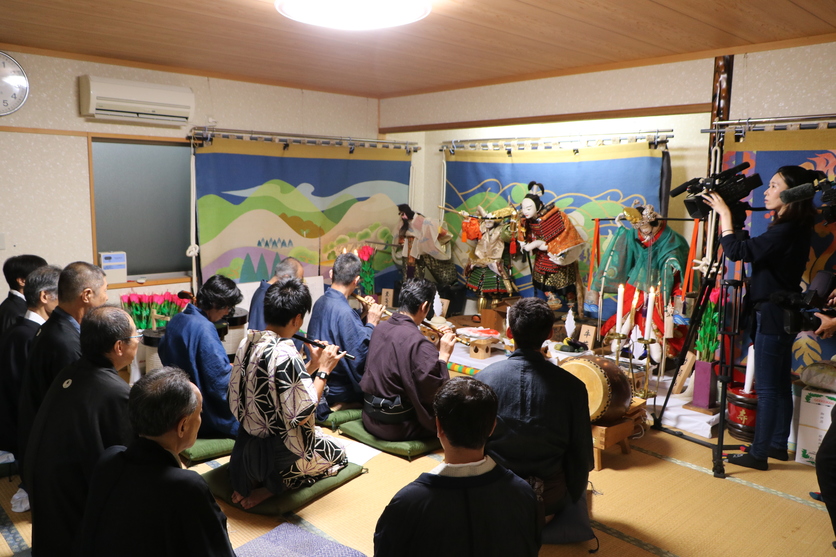 和装姿の人形に向かって正座をして笛を吹いている人たちの写真