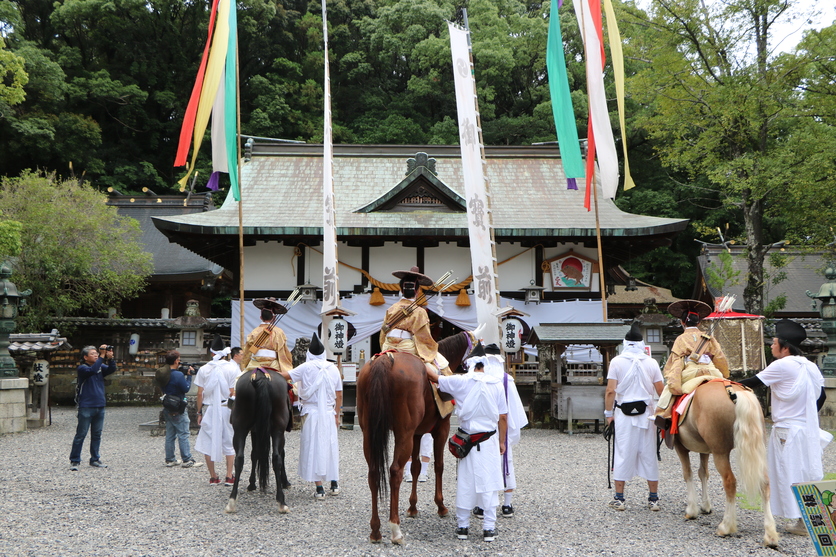 社殿に向かって立つ馬に乗った3人とそのわきに立つ白い装束を着た人たちの写真