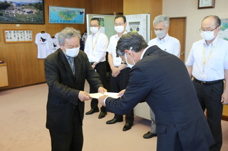 白いワイシャツ姿の男性が4人並び、その前で報告書の受け渡しをしている二人の男性の写真