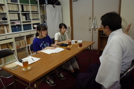 白衣姿の男性が机を挟んで向かい側に座っている人達に話をしている写真