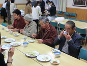 食堂でテーブルに並んで着席した人々が料理を食べている写真