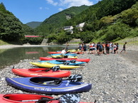 川辺の砂利浜に並べられた複数のカヤックと、奥で集まって談笑している人々の写真