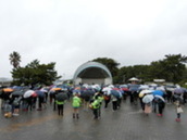 半円形の屋外ステージ前に、傘を差した人々が集まっている写真