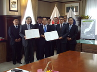 スーツ姿の男性達が並んで立ち、前2人が書類を持っている写真