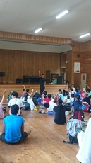 体育館で床に座った多くの人が前方の男性に向かって手を挙げている写真