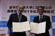 市のロゴ入りボードの前で書類を持った男性2人が並んでいる写真