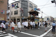 街中の交差点を白い法被を着た人々が山車を曳いて渡っている写真