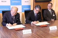 3人の男性が座り、内2人が手元の書類に署名している写真