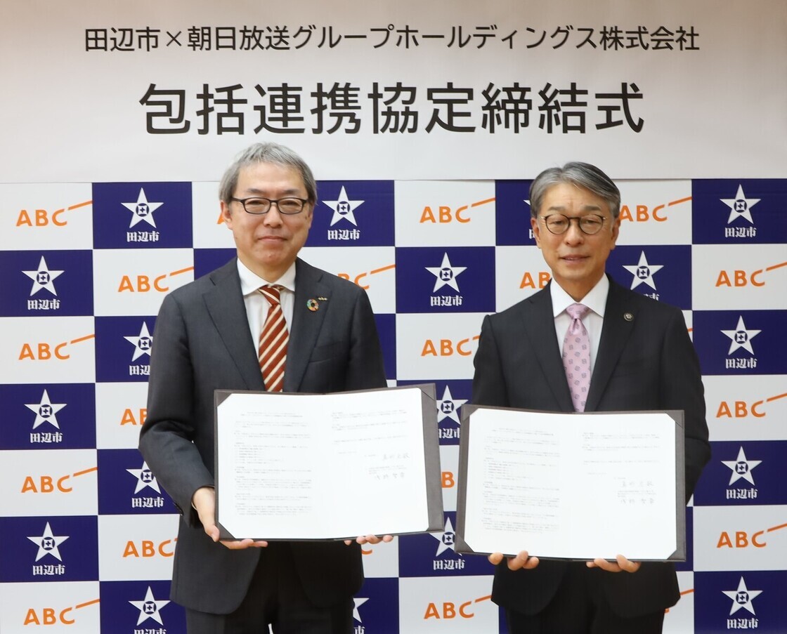 市のロゴ入りボードの前で男性二人が協定書を見せながら横に並んでいる写真