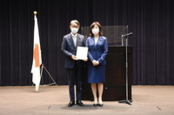 日本国旗が置かれた壇上で、紙を持った男性とブルーのスーツを着た女性が並んでいる写真