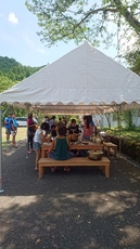 屋外に設置されたテントの下で、人々が木製ベンチに集まって談笑している写真