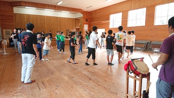 木造の広い室内で多くの人が輪になっており、右手前には小太鼓を叩く男性が立っている写真