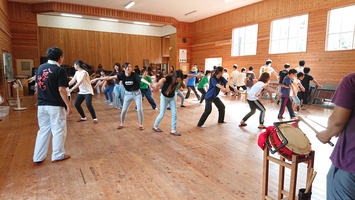 木造の広い室内で多くの人が輪になっており、全員が輪の外側に向かって両手を広げている写真