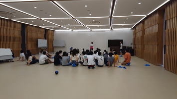 広い室内で床に座った多くの人が前方で立っている人の話を聞いている写真