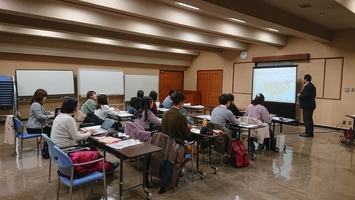 机に着席している複数の人と前方でスクリーンを使って説明している男性の写真