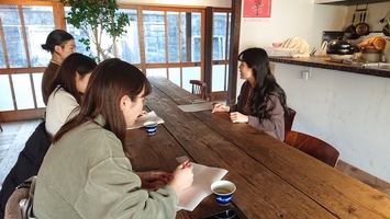 木のテーブルの右手側に座った女性が向かい側の3人へ話をしている写真