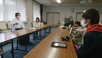 長机の片側に複数の人が着席し、向かい側にいるマスクを着けた男性の説明を聞いている写真