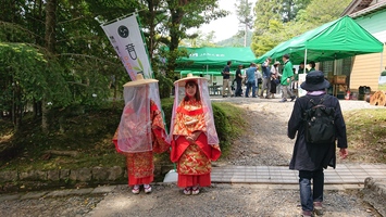 2人の赤い衣装を着た人が並んで立ち、その奥のテントでは人々が集まって談笑している写真