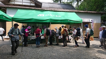 屋外のテント前に、帽子やリュックを身につけた多くの人が並んで順番に手続きをしている写真