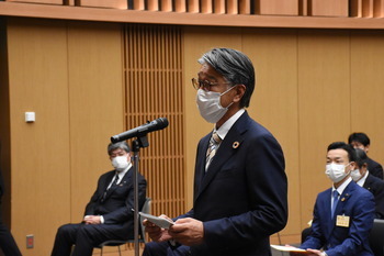 スタンドマイクの前で、マスクをして眼鏡をかけた男性が立っている姿を斜め右から写した写真