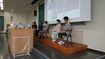 背後にスクリーンのある壇上で5人の男女が椅子に座っている写真