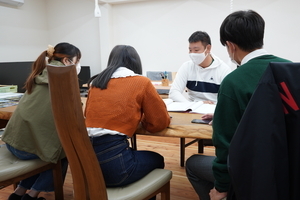 資料をもとに説明している男性とそれを聞く3人の男女の写真