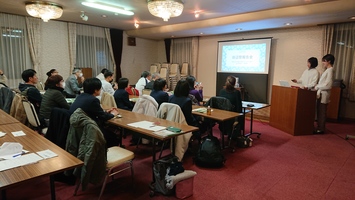 会議室で複数の人が机に着席し、前方では2人がスクリーンを使って発表している写真