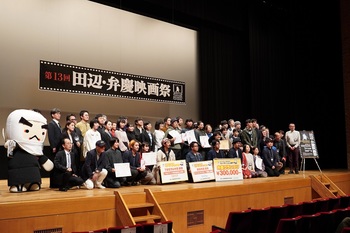 壇上で、たくさんの男女と弁慶のキャラクターがスクリーンを背景に並んでいる写真