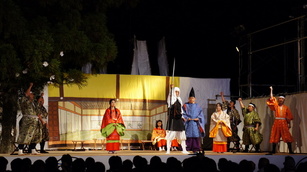 舞台上で、白袈裟を被った弁慶役の男性が右手を高く上げ、その周りを囲んでいる武士の恰好をした男性達も右のこぶしを上げている姿と観客の背後が写っている写真