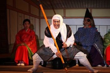 赤い服を着た女性と青い服の眼帯をした男性の前で、棒を持った弁慶の格好をした人が腰をかがめて足を大きく開いて力強く叫んでいる写真