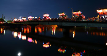 橋の上を進む複数の笠鉾の明かりが水面に反射している写真