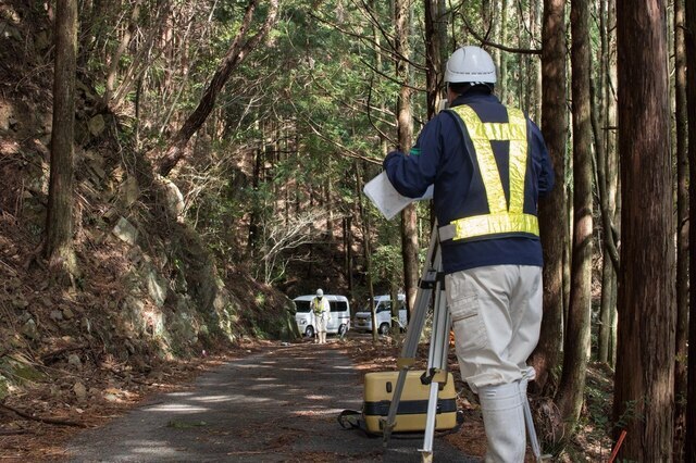 山の中にある舗装路で測量を行う作業着姿の人の写真