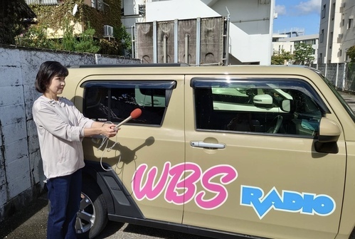 「WBS」と書かれたステッカーの貼られた車の横でマイクを前に出している女性の写真