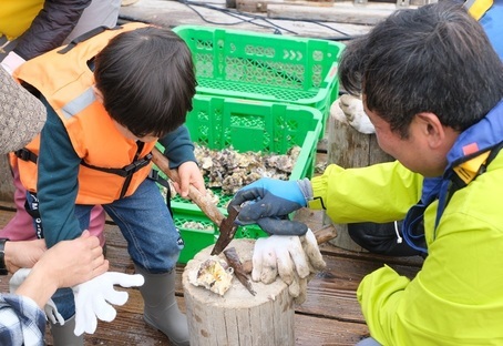 ライフジャケットを着た男の子が男性に教わりながら牡蠣にハンマーを向けている写真