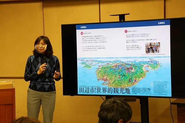 資料が映ったテレビの横でマイクを持った女性が立っている写真