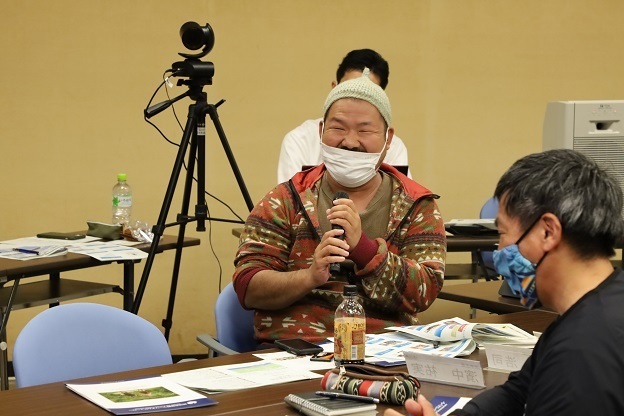 テーブルに着いている男性がマイクを持って話をしている写真