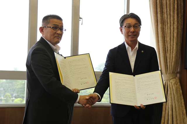 男性二人が片手で見開きの書類を持ち、もう片方の手で握手している写真