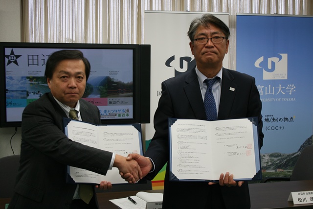 スーツ姿の男性二人が片手で見開きの書類を持ち、反対の手で握手している写真