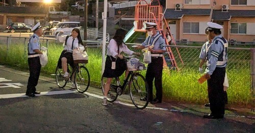 夕方の道路脇で自転車に乗った学生たちに警察官が交通安全グッズを配布する写真