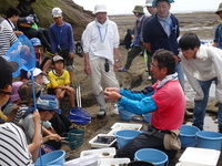 砂地でバケツに囲まれた男性が前に差し出した手を真剣に見つめている人たちの写真