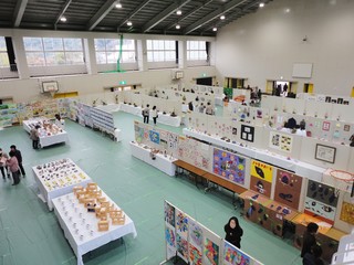体育館で白いパネルや机に絵画や工作などの作品が並んでいる写真